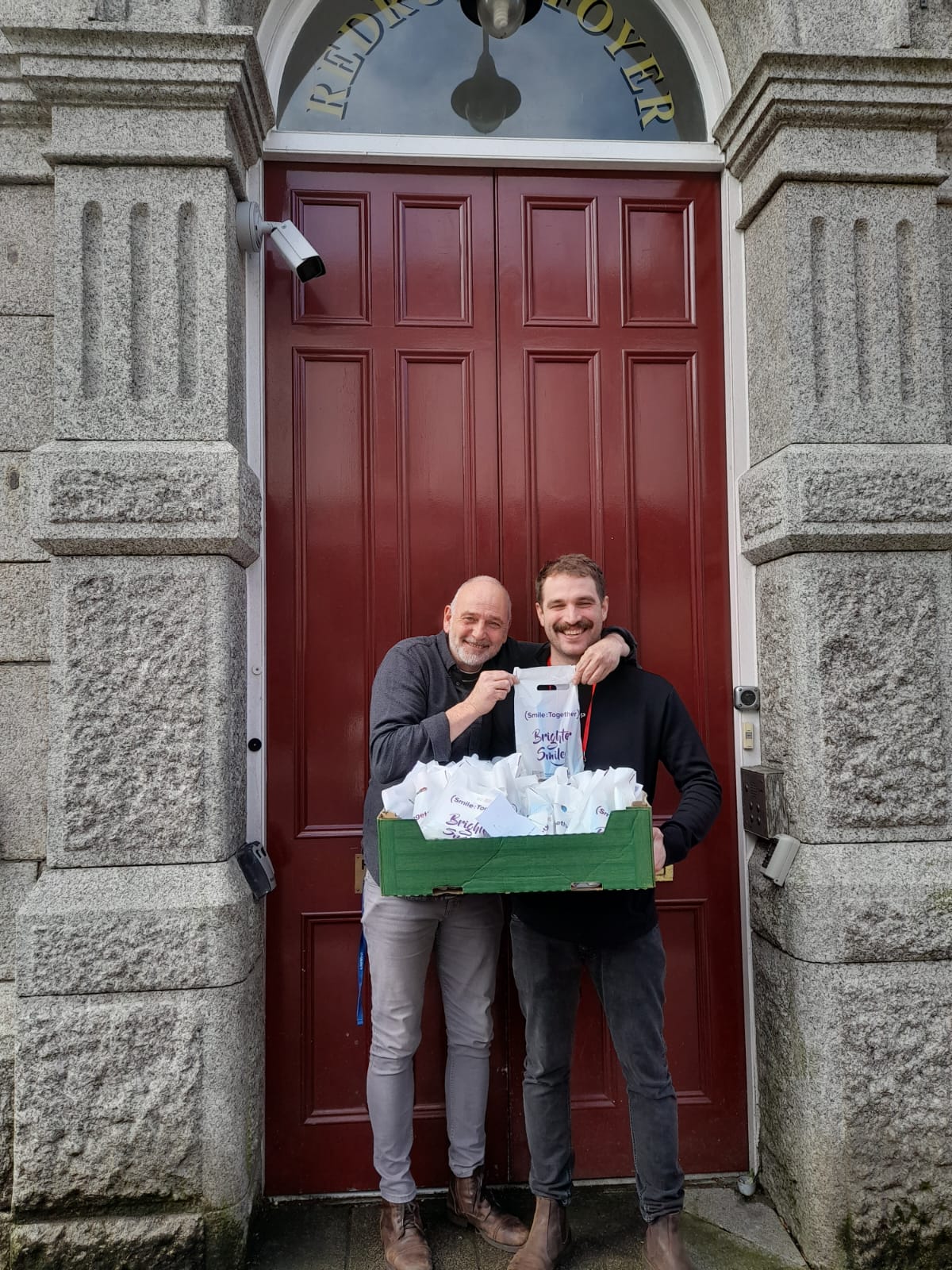 two men smiling and holding oral health packs outside of Redruth Foyer