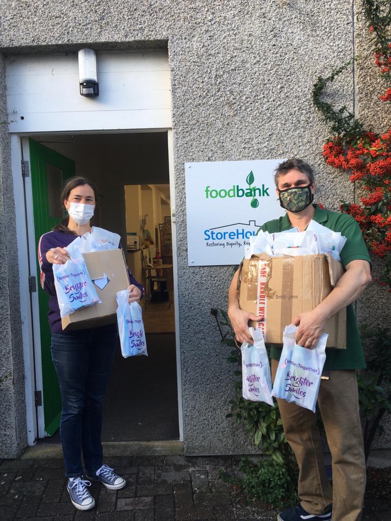 Man and woman masked standing outside foodbank holding boxes of oral health packs