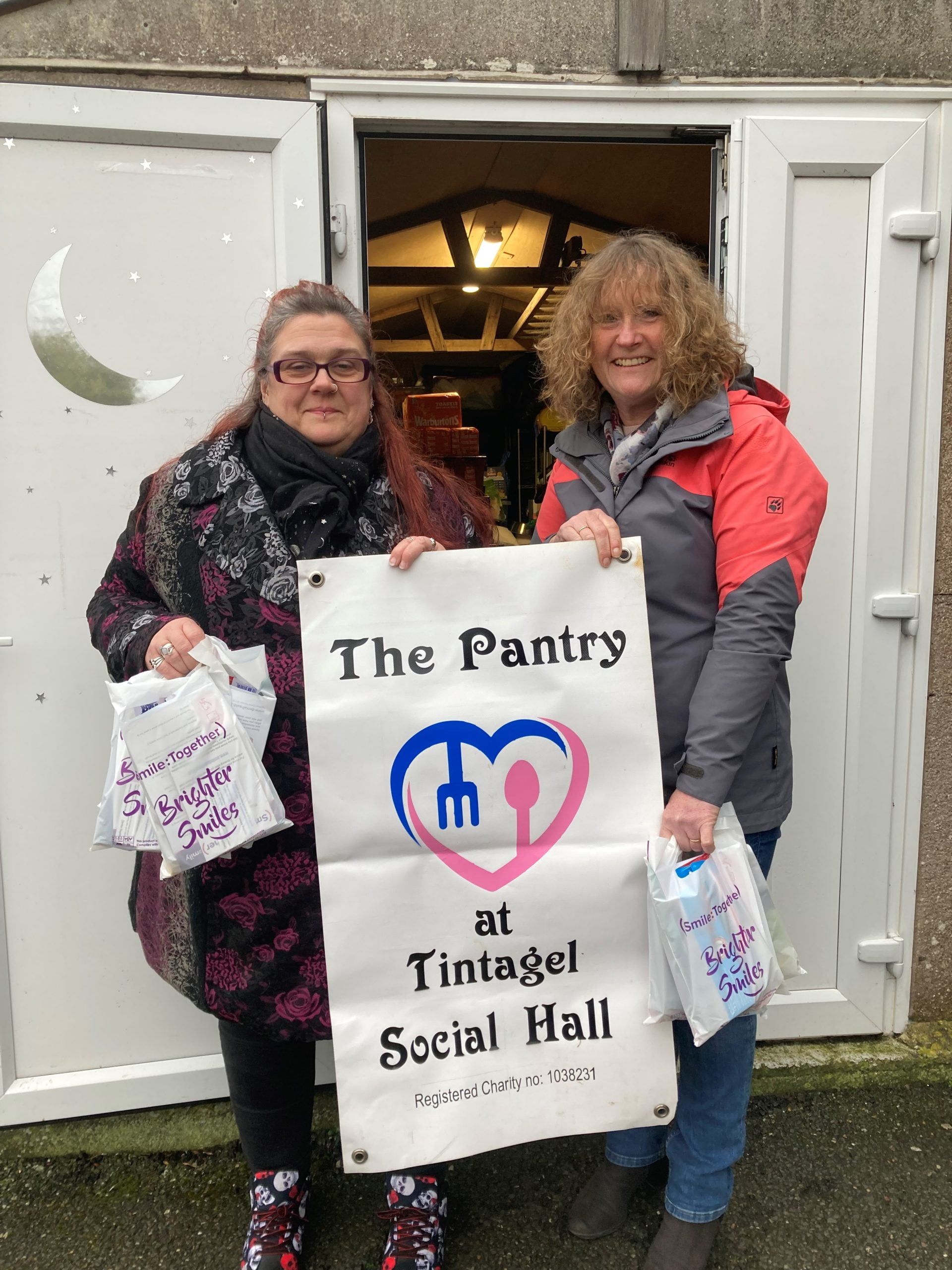 two women holding oral packs and large sign reading 'the Pantry at Tintagel social hall. registered charity.'