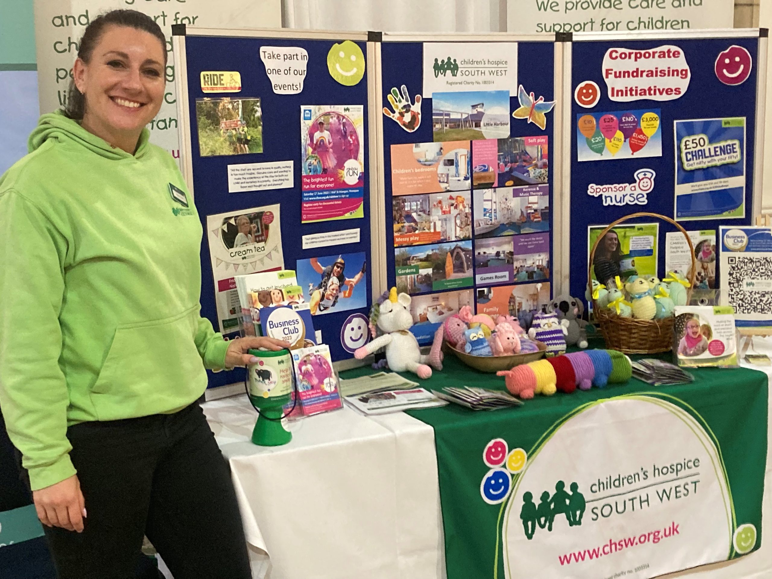 Amanda from Children's Hospice South West in green hoodie stood by her stall with money pot, posters, knitted toys