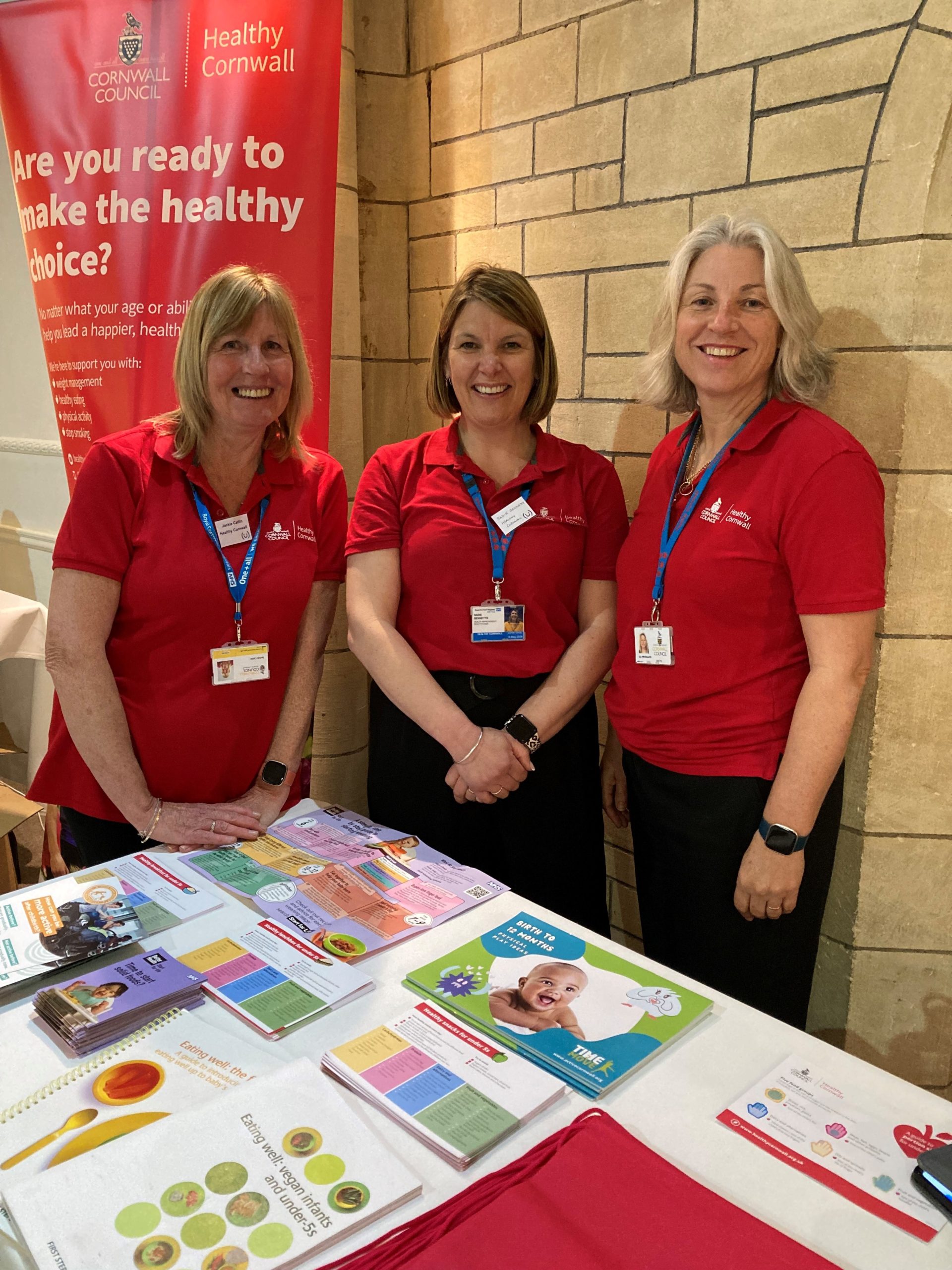 three ladies from healthy cornwall in red tops stood by their stand behind poster reading 'are you ready to make the healthy choice'